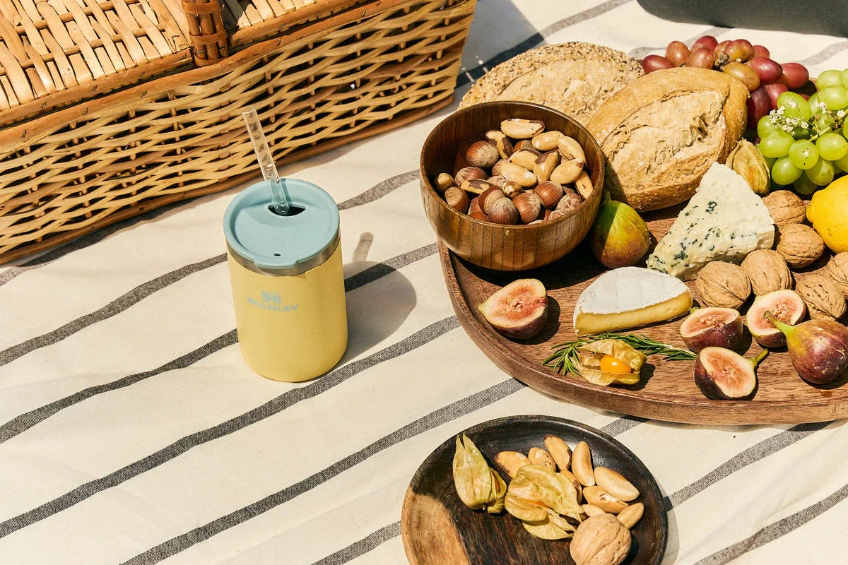 Um Can Cooler Stanley perto de uma cesta e comidas, representando que está acontecendo um piquenique.