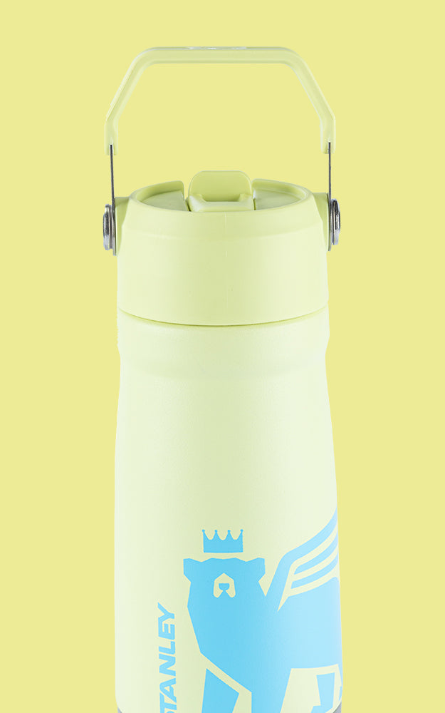 Garrafa térmica de cor amarela clara com uma alça de metal na parte superior. A garrafa possui um design minimalista com um logotipo azul claro de um urso com uma coroa e asas. A palavra "STANLEY" está escrita verticalmente ao lado do urso. O fundo da imagem é da mesma cor amarela clara da garrafa, criando um efeito de continuidade visual.